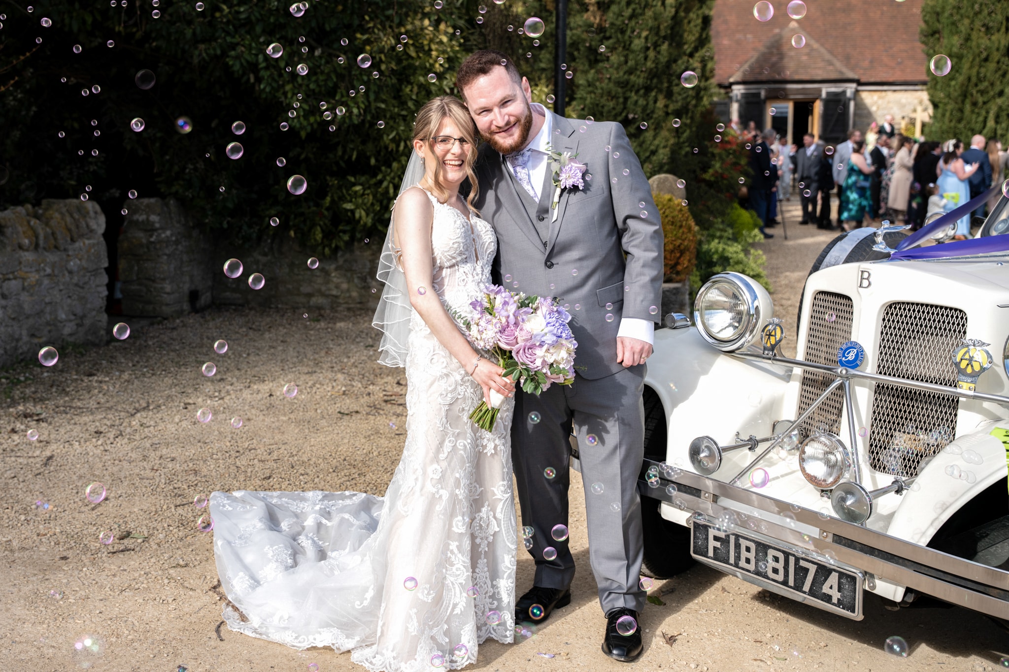 Bride and groom laughing together beside vintage wedding car with bubbles at Notley Tythe Barn