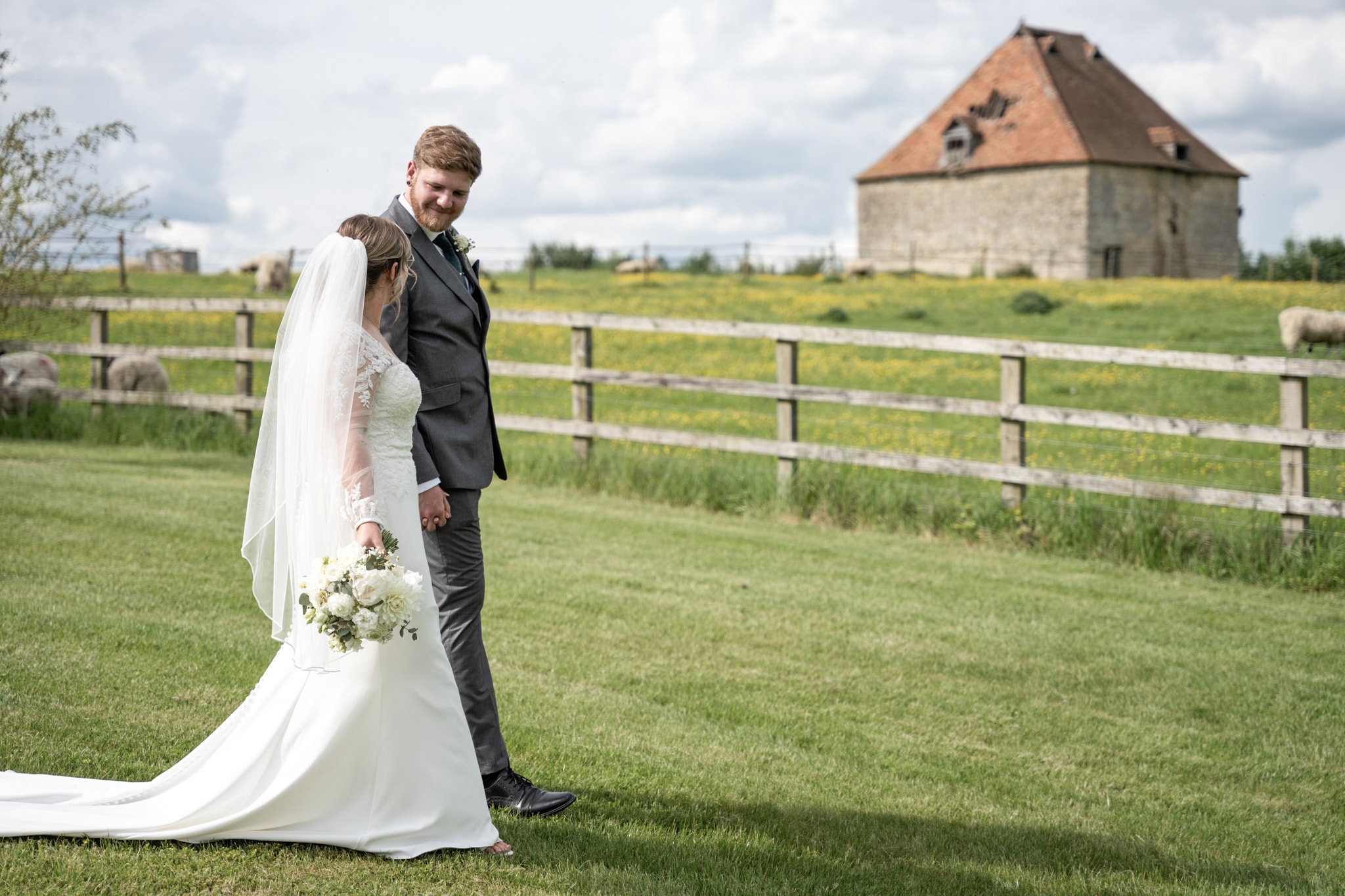 Bride and groom holding hands in meadow with historic stone dovecote and sheep in background at Notley Tythe Barn