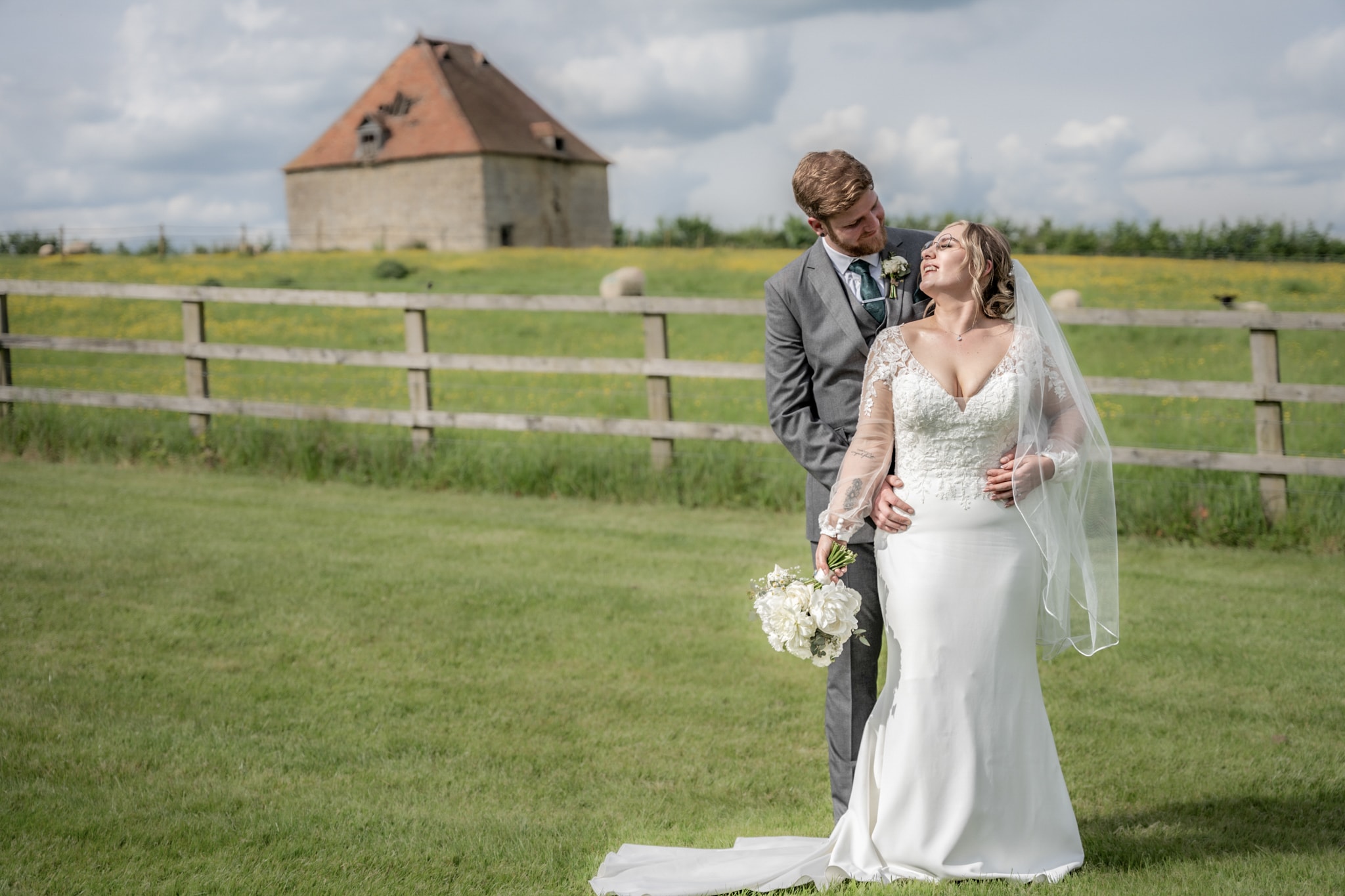 Groom embracing bride from behind as she laughs in meadow with stone dovecote backdrop at Notley Tythe Barn