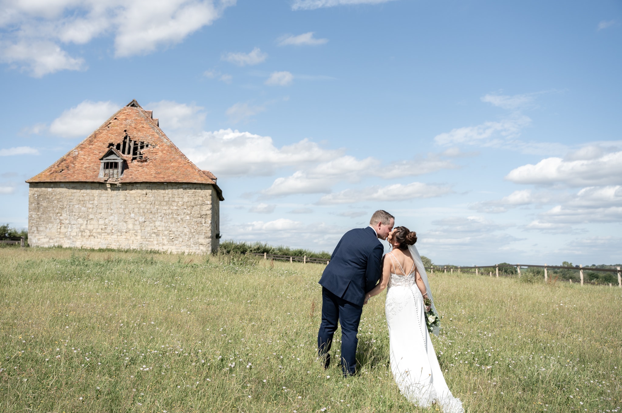 Bride and groom kissing in wildflower meadow with rustic stone building and blue sky at Notley Tythe Barn