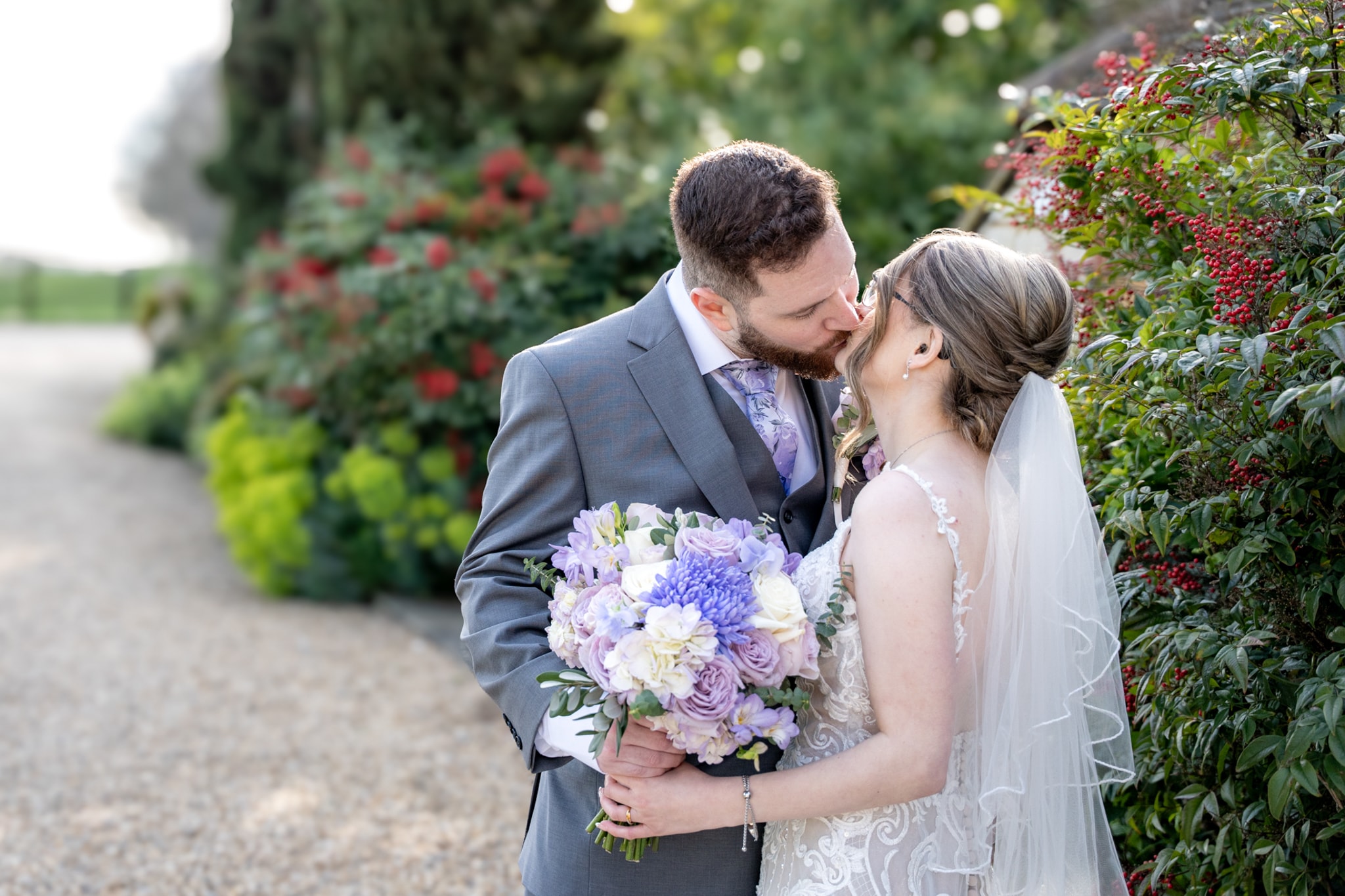 Bride and groom kissing beside garden path with purple and white bouquet and red berry hedgerow at Notley Tythe Barn