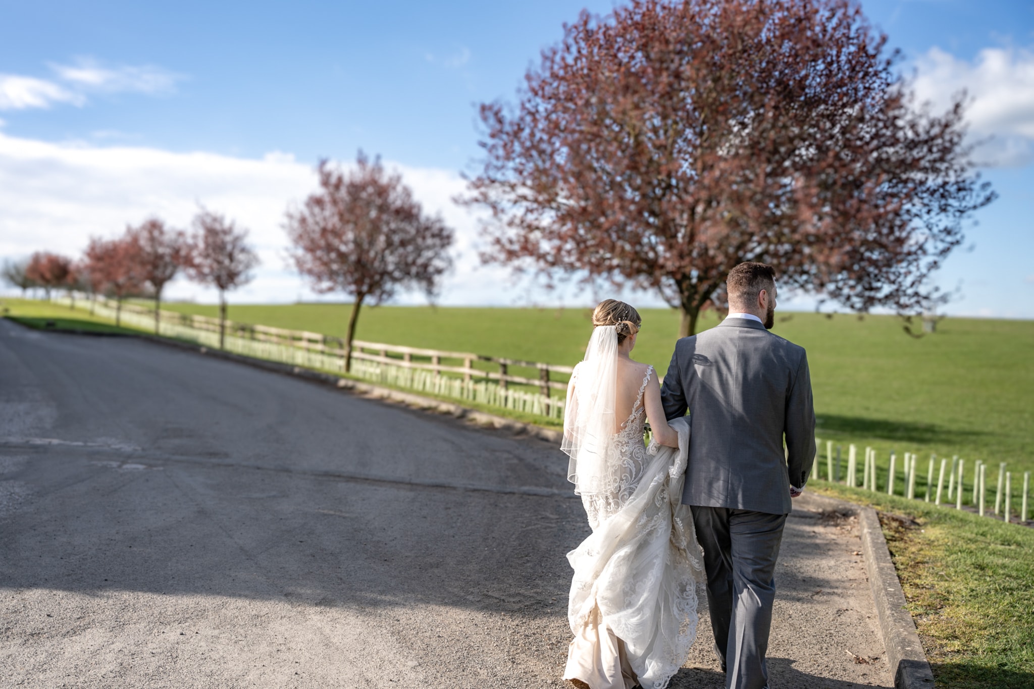 Bride and groom walking away down tree-lined driveway with blossom trees and open countryside at Notley Tythe Barn