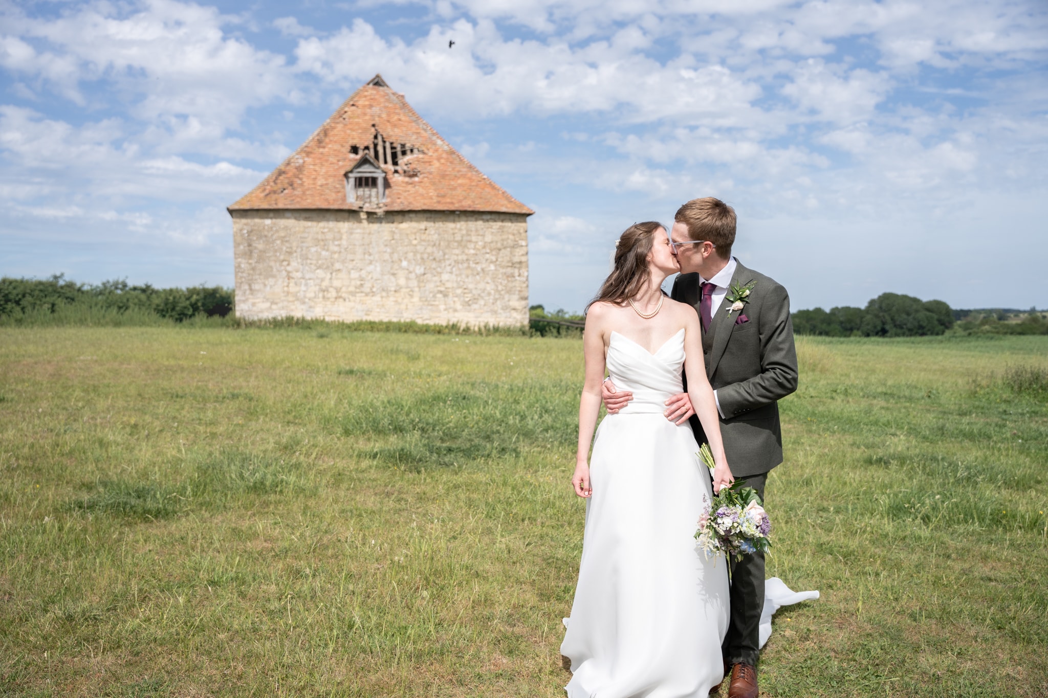 Bride and groom kissing in open meadow with rustic stone dovecote and blue sky at Notley Tythe Barn
