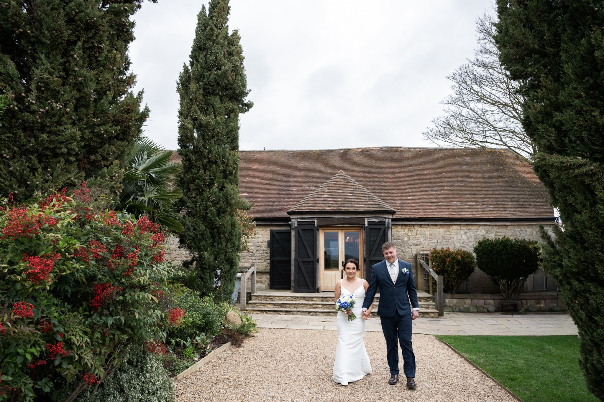 Bride and groom walking hand in hand along gravel path with tall cypress trees and stone barn at Notley Tythe Barn
