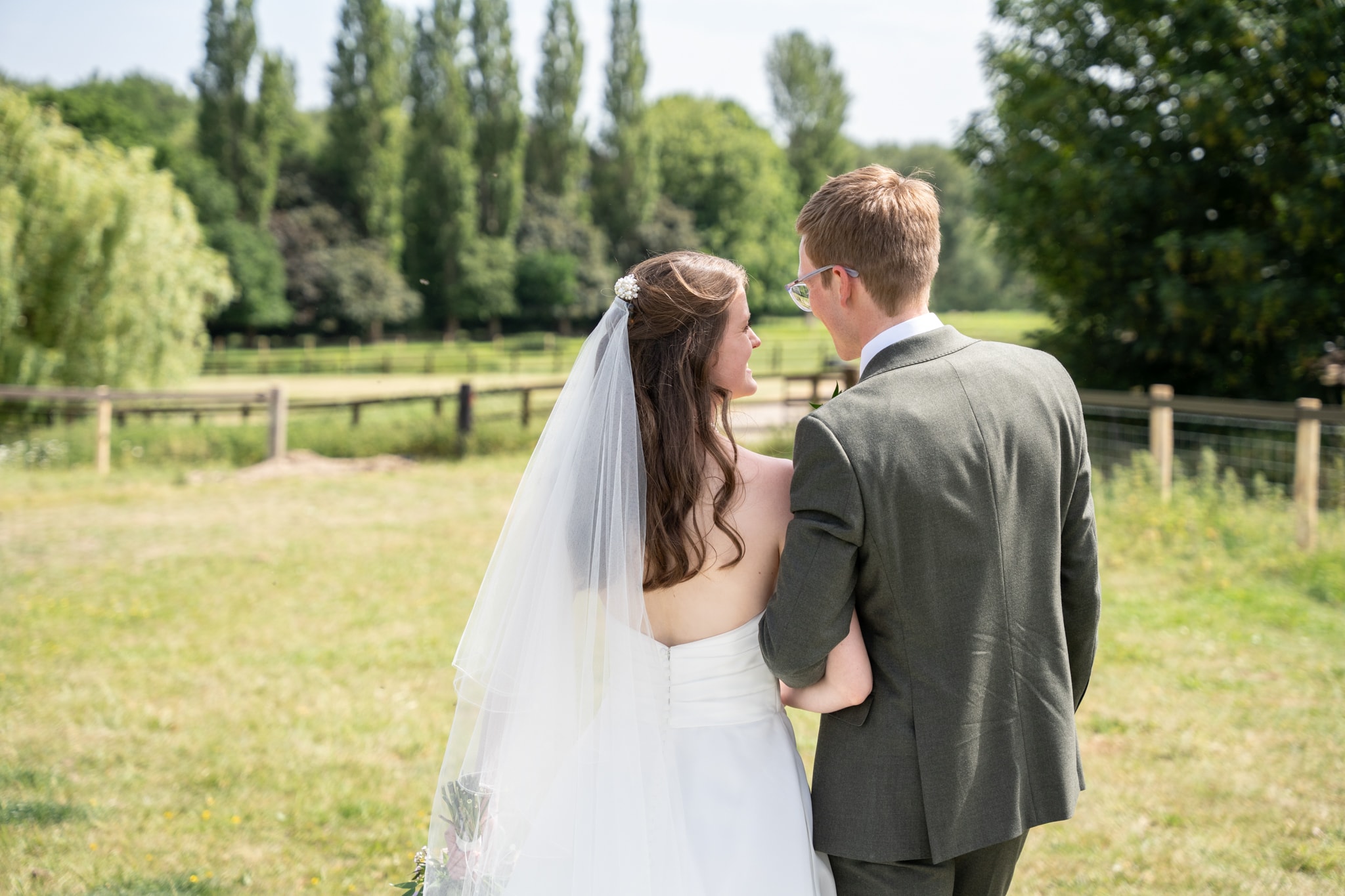 Bride and groom from behind arm in arm looking out over sunny meadow with willow trees at Notley Tythe Barn