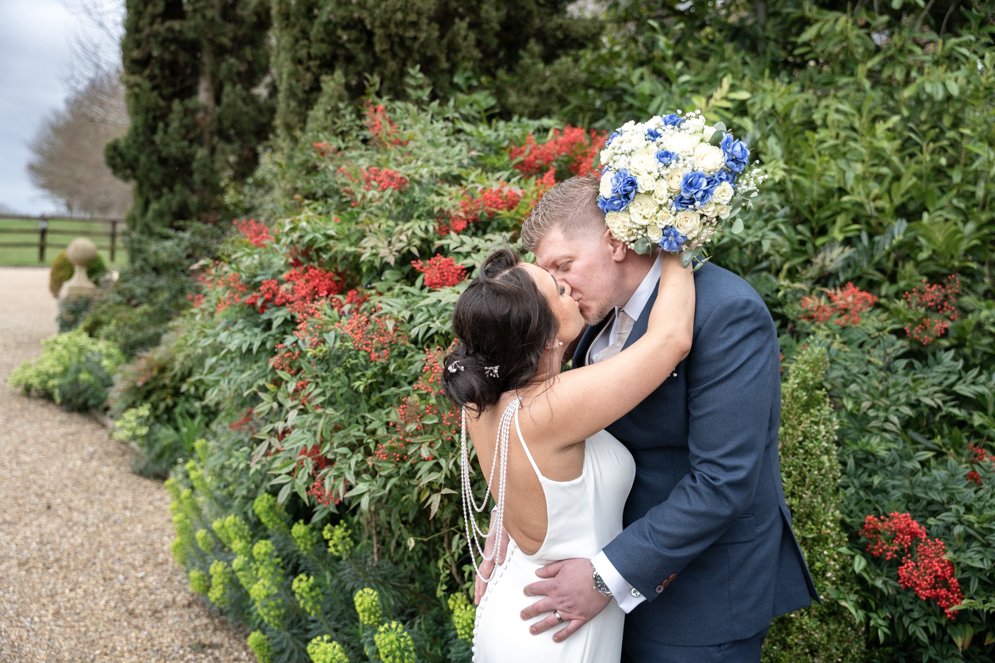 Bride and groom kissing in front of red berry hedgerow with blue and white bouquet at Notley Tythe Barn
