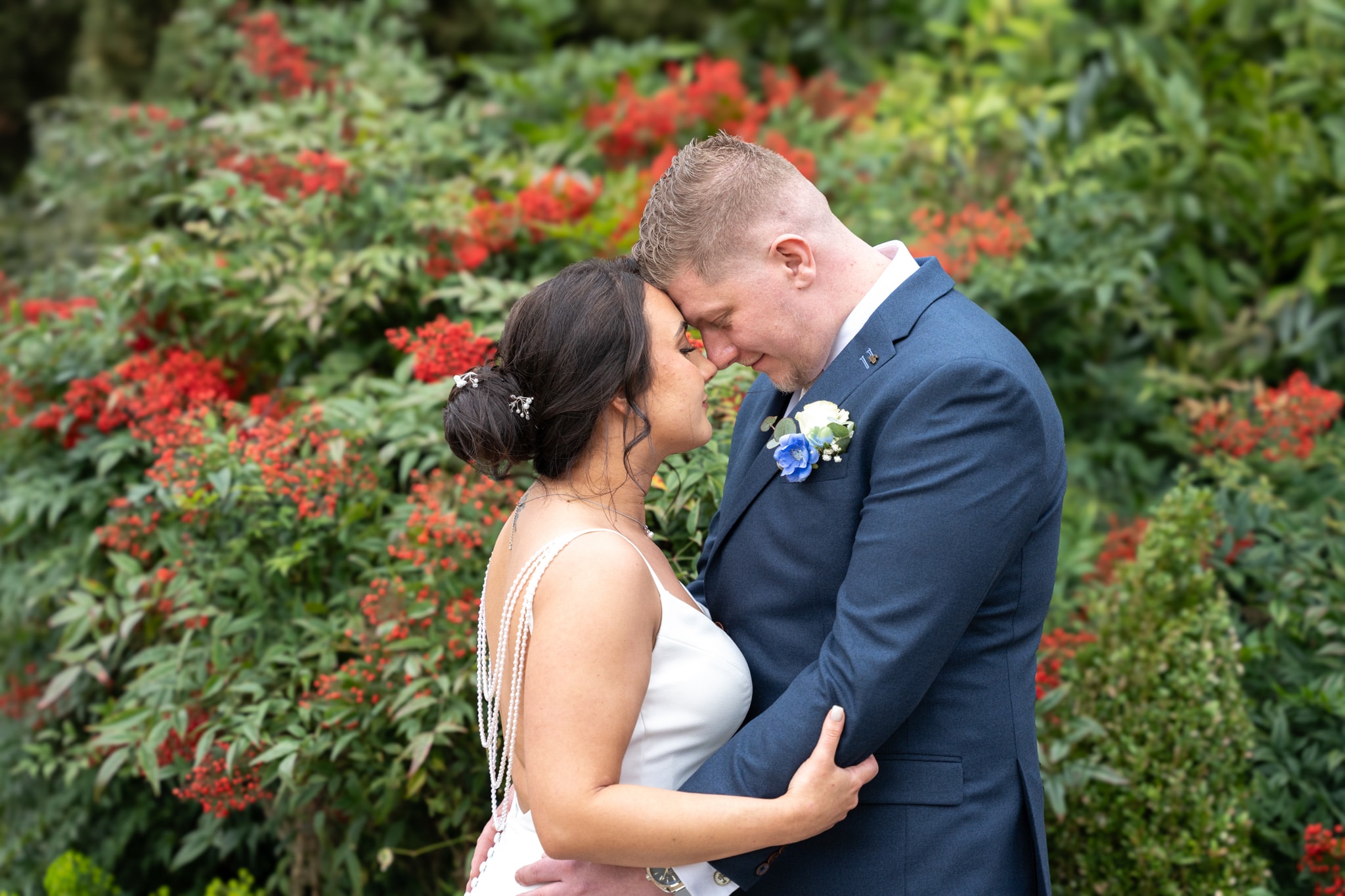 Bride and groom forehead to forehead embracing in front of vivid red berry hedgerow at Notley Tythe Barn