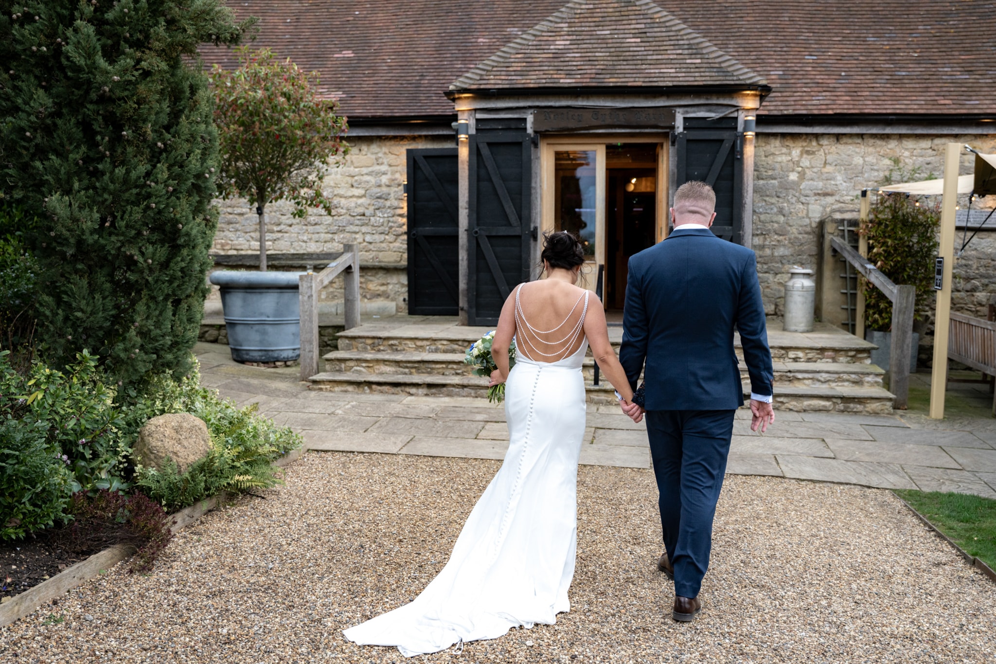 Bride and groom walking away hand in hand toward stone barn entrance at Notley Tythe Barn