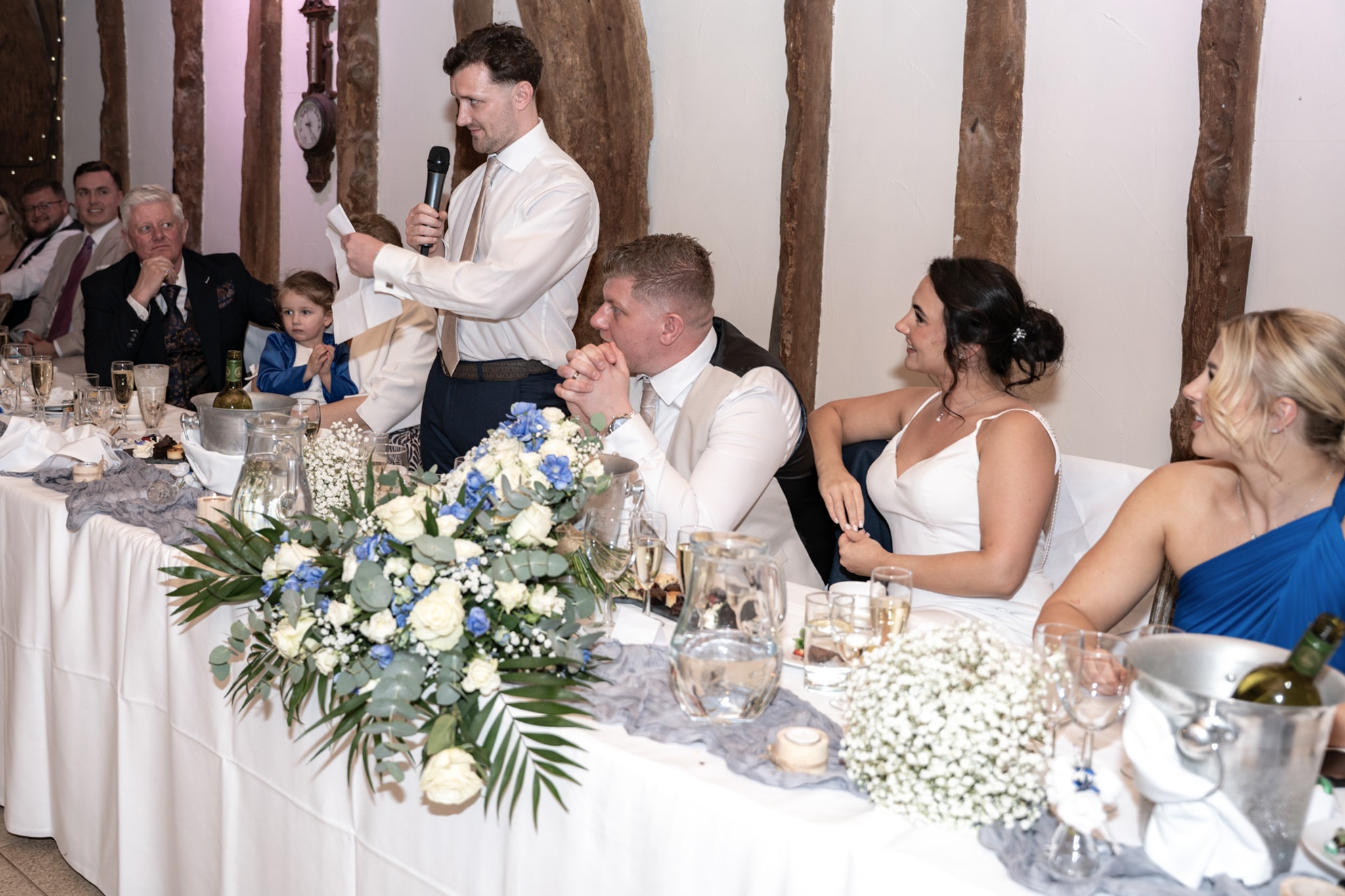 Best man giving speech at top table with bride and groom smiling and blue floral centrepiece at Notley Tythe Barn