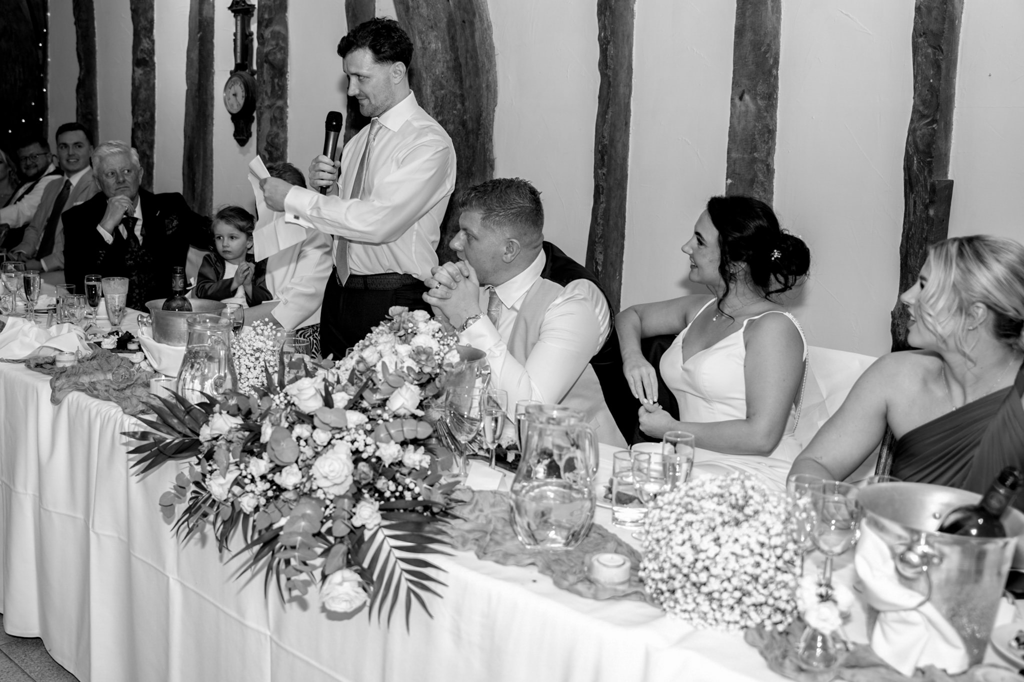 Black and white image of best man speech with bride and groom at top table inside Notley Tythe Barn