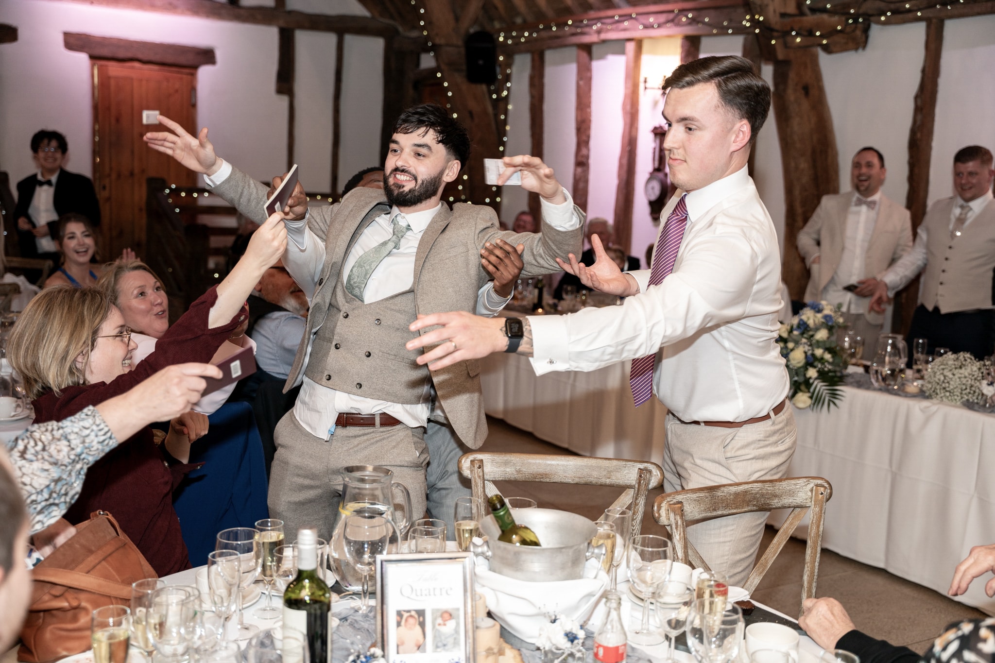 Wedding guests celebrating with cards at dinner table inside Notley Tythe Barn reception room with fairy lights and oak beams