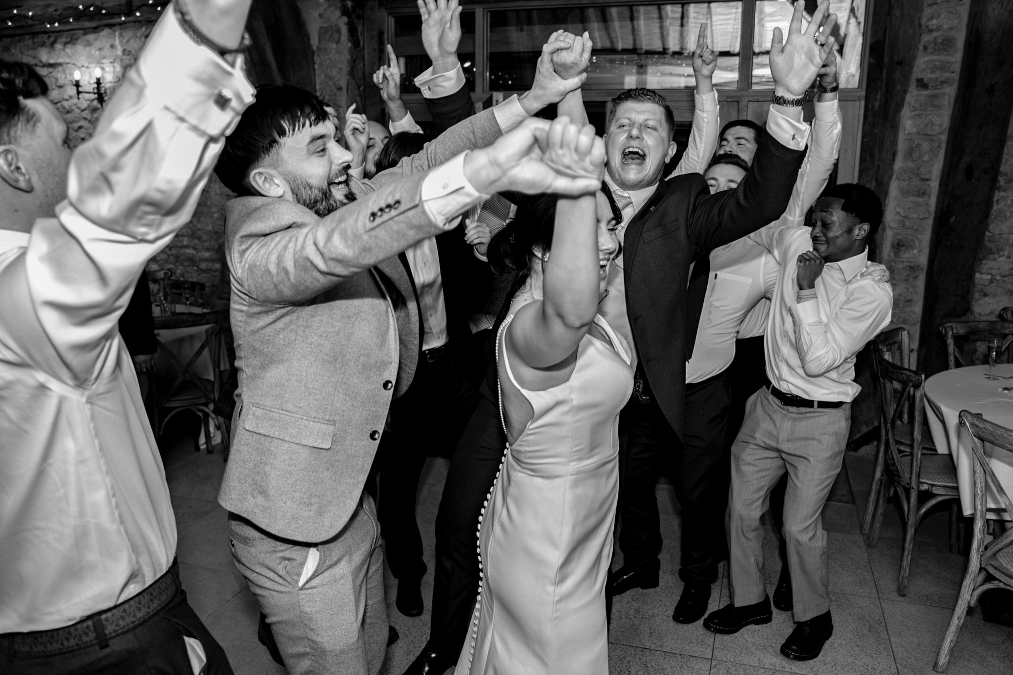 Bride and wedding guests celebrating with arms raised on dance floor at Notley Tythe Barn reception in black and white